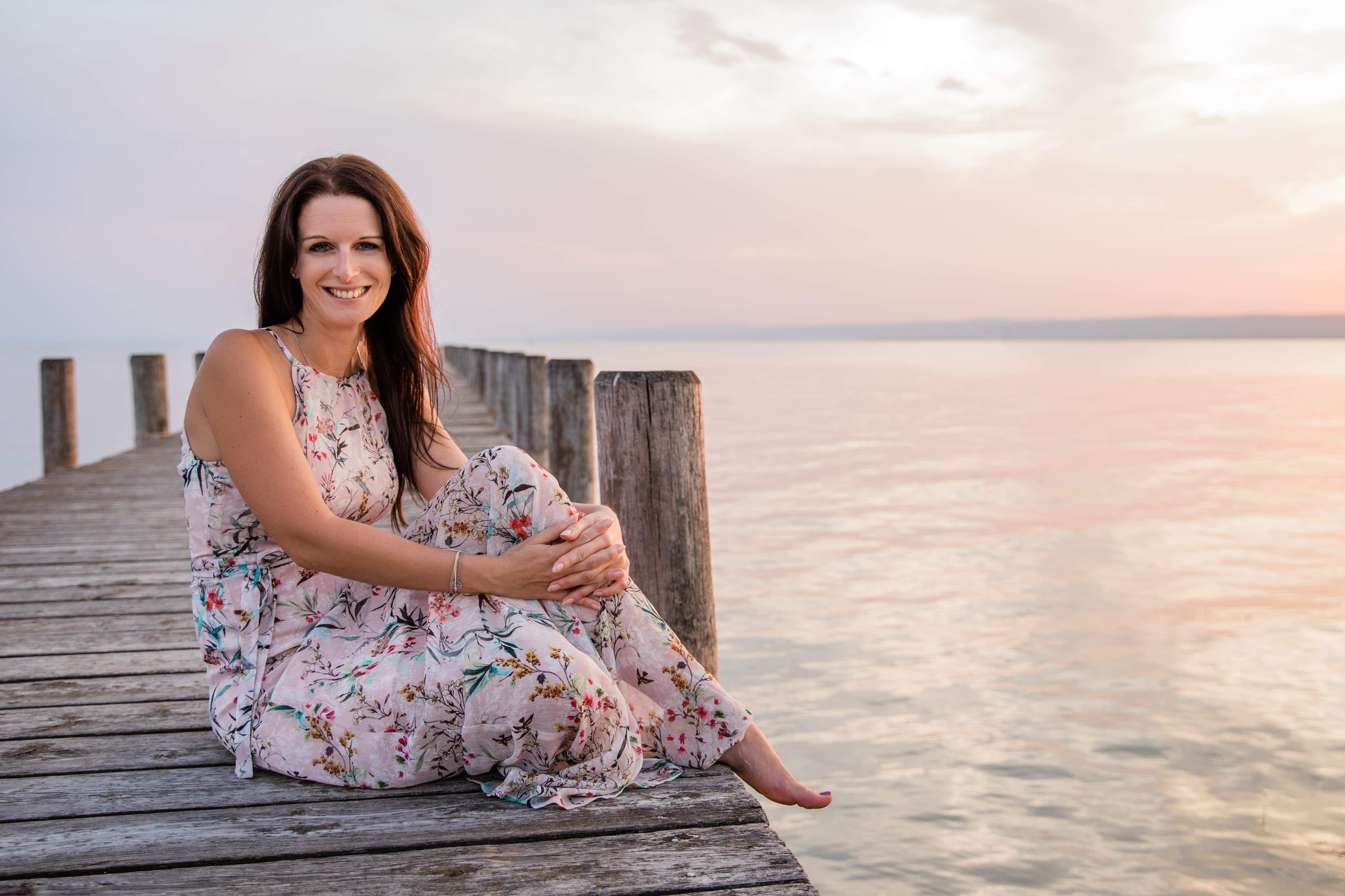 Angelika sitzt lächelnd auf einem Steg am See bei Sonnenuntergang